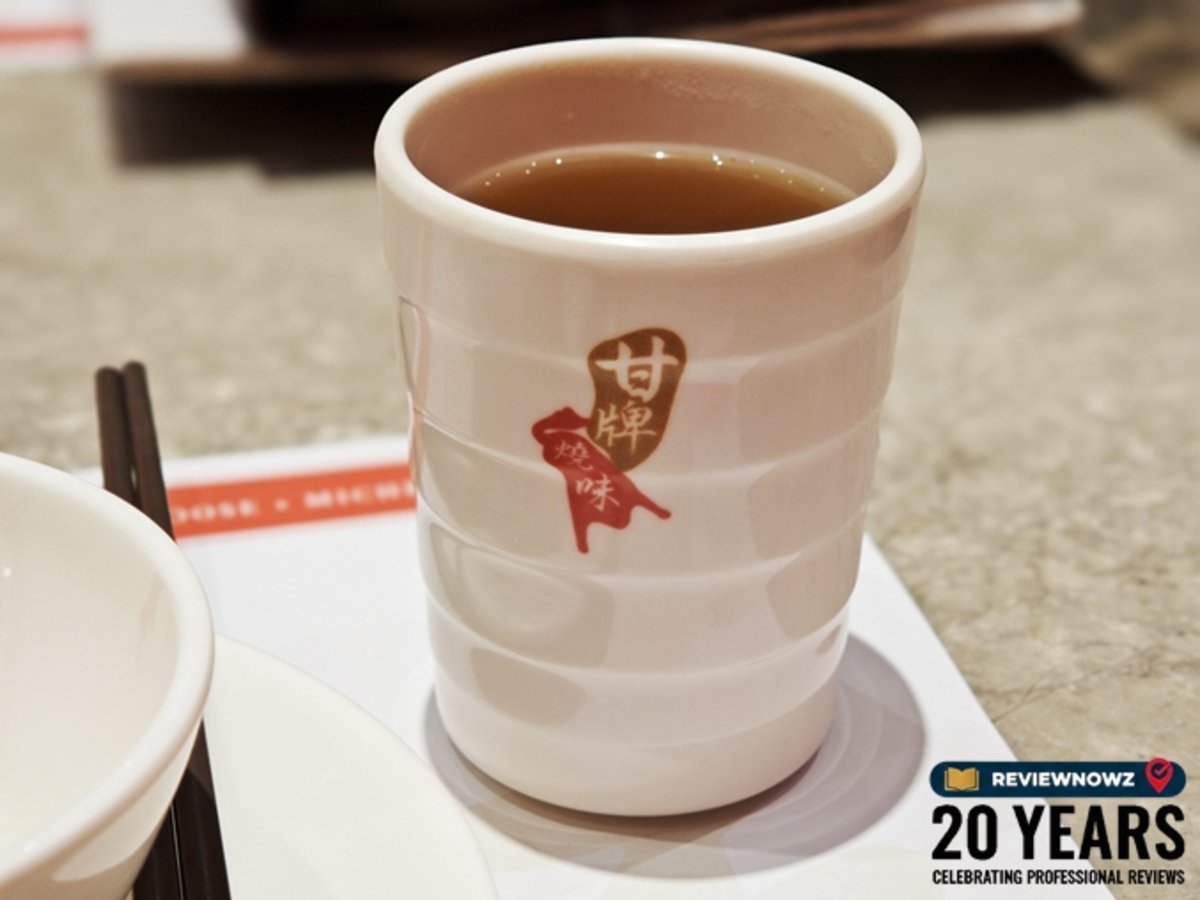 แก้วชาเซรามิกสีขาวพิมพ์โลโก้ร้าน Kam's Roast บรรจุน้ำชาจีนร้อนสีเข้มข้น วางเคียงคู่กับตะเกียบบนโต๊ะอาหาร