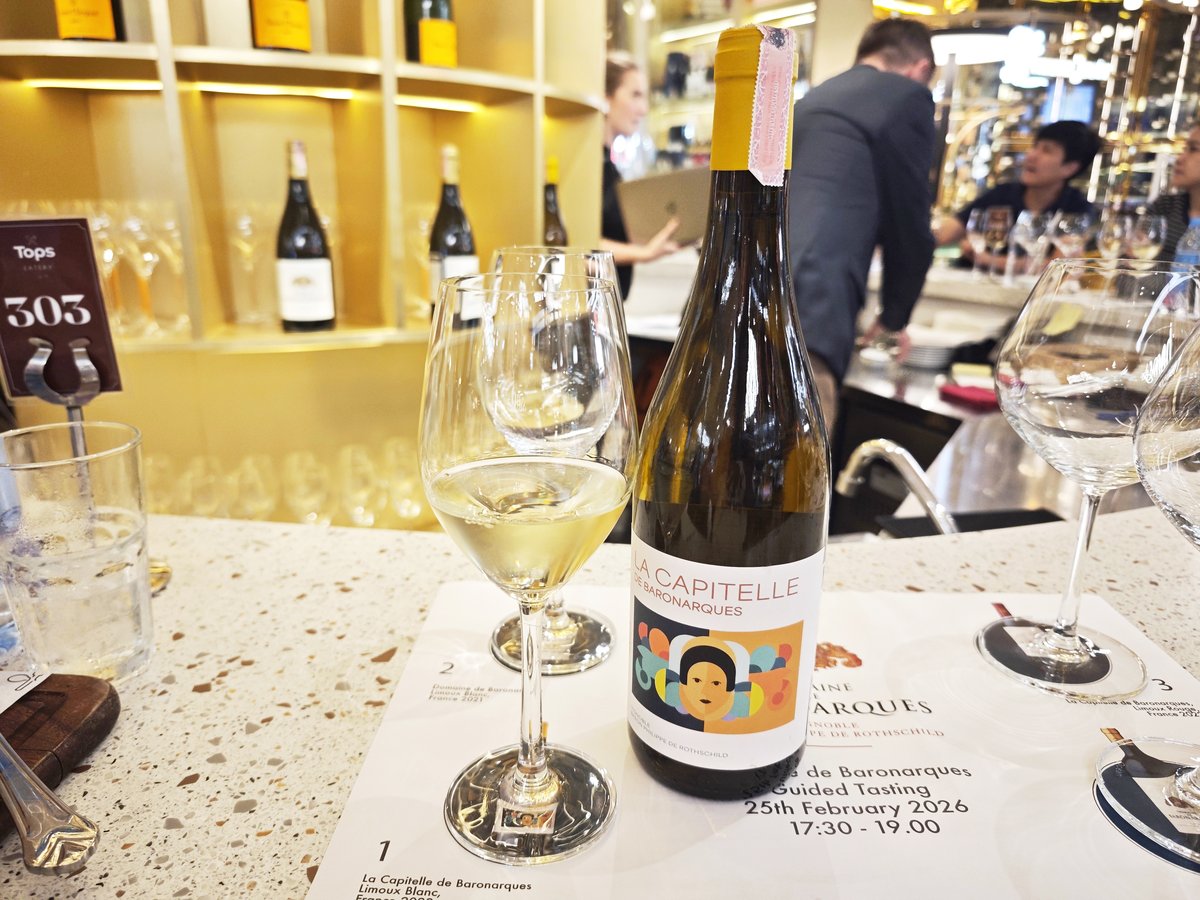 ขวดไวน์ขาว La Capitelle de Baronarques Blanc ดีไซน์ฉลากรูปหน้าคนสีสันสดใส วางคู่กับแก้วไวน์ที่มีน้ำไวน์สีเหลืองทองอ่อนๆ บนแผ่นรอง Tasting Mat ที่เคาน์เตอร์บาร์ Tops Central Park รีวิวโดย ReviewNowz