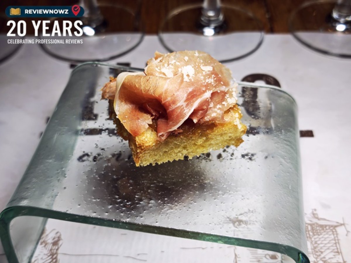 เมนู Focaccia, San Daniele prosciutto, onion jam ขนมปังโฟกาเซียท็อปด้วยพาร์มาแฮมสไลด์บางและแยมหอมใหญ่ โรยหน้าด้วยพาร์เมซานชีส จัดเสิร์ฟอย่างหรูหราบนแท่นกระจกใสโค้ง บนกระดาษรองจานสำหรับงานรีวิวอาหาร ณ ห้องอาหาร Attico โรงแรม Radisson Blu Plaza Bangkok