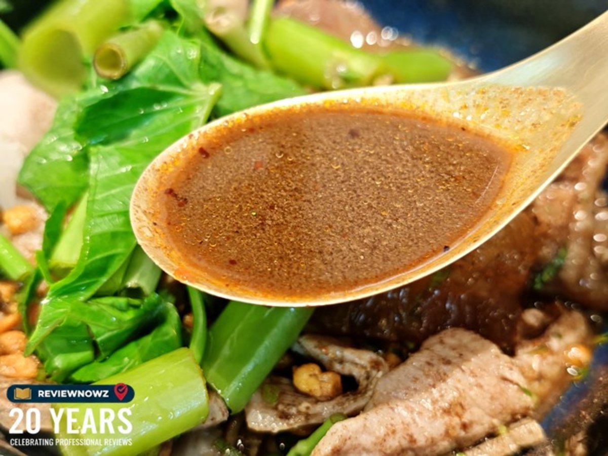 ช้อนทองเหลืองตักน้ำซุปก๋วยเตี๋ยวเรือน้ำตกสีเข้มข้น รายล้อมด้วยผักบุ้งสดและเครื่องก๋วยเตี๋ยวเรือ ร้านก๋วยเตี๋ยวเรือทองสมิทธ์ สาขาพาร์คสีลม ReviewNowz