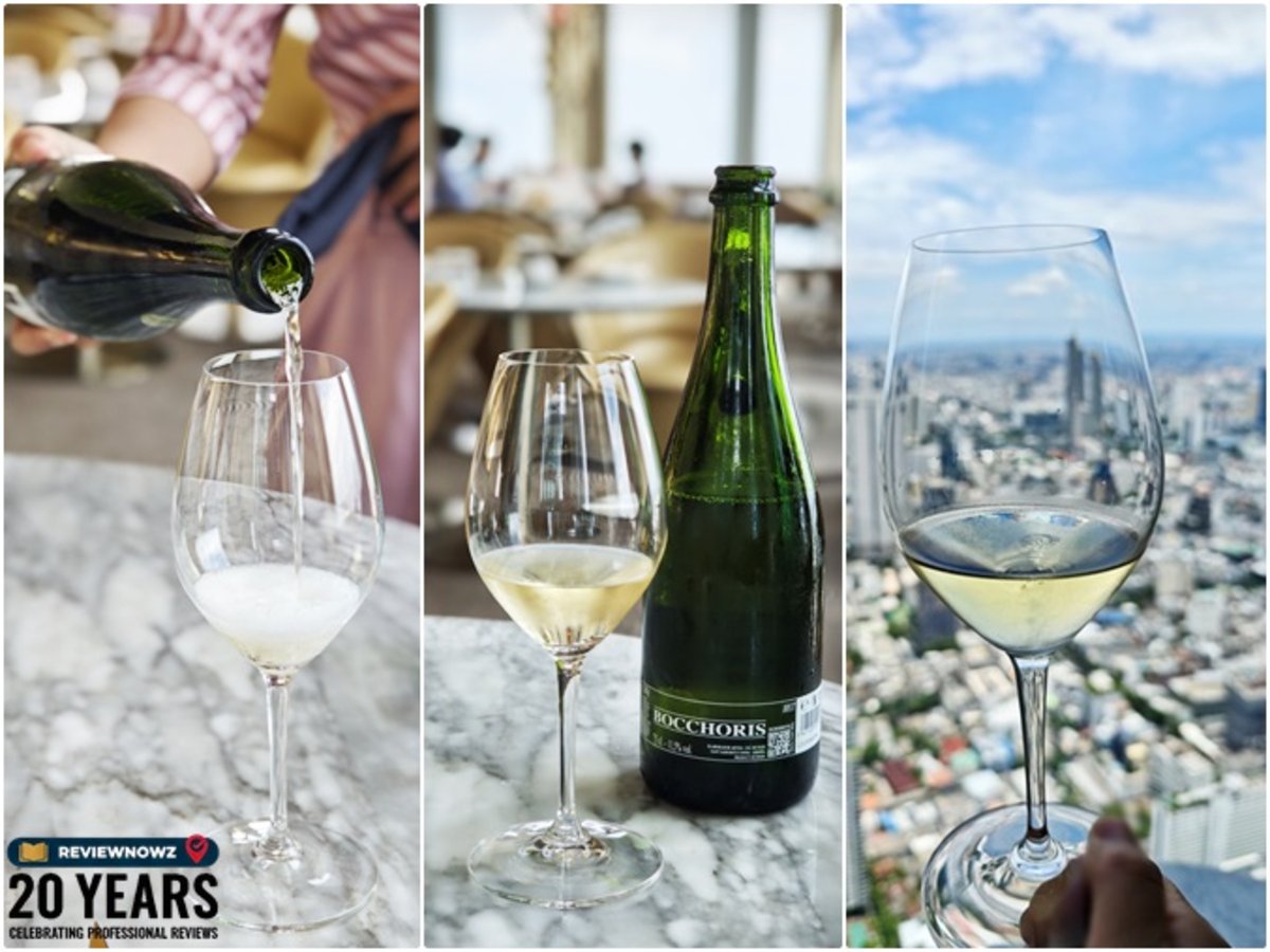 การรินสปาร์กลิ้งไวน์ขาว Bocchoris Cava Brut ลงในแก้วทรงสูง วางคู่ขวดไวน์และแก้วไวน์บนโต๊ะหินอ่อนพร้อมวิวท้องฟ้ากรุงเทพฯ มุมสูง ร้าน OJO Bangkok ชั้น 76 โรงแรม The Standard, Bangkok Mahanakhon ReviewNowz